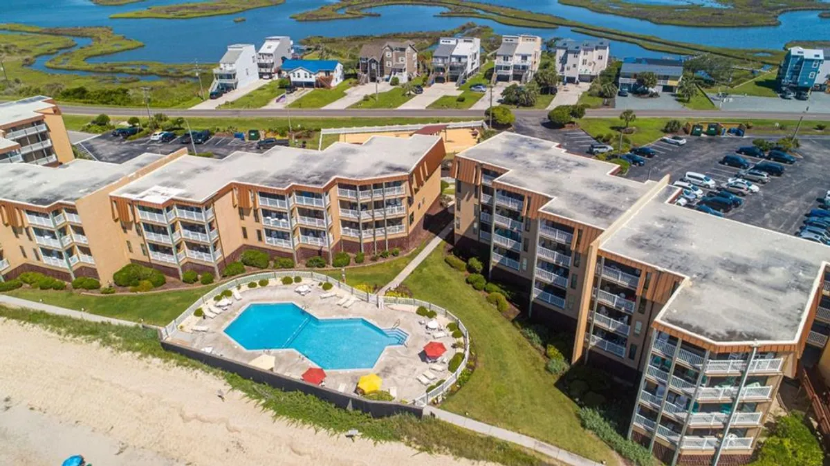 A bright exterior scene featuring the oceanfront swimming pool framed by lush greenery and a few nearby homes, with families swimming, sunbathing, and gathering poolside. The beachfront setting and vibrant activity encapsulate the resort's lively, vacation-like atmosphere.