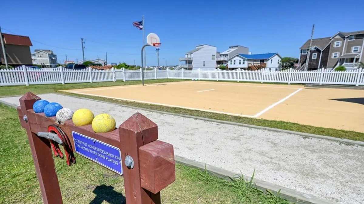 An active exterior scene featuring a basketball court and bocce balls, surrounded by cheerful summer greenery and a white picket fence, with families and friends enjoying outdoor games together.