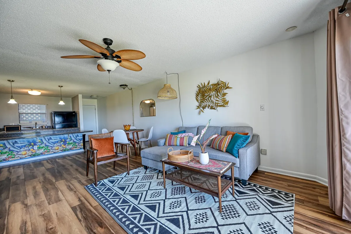 This bright, open-plan living space features a modular sofa adorned with colorful cushions, a dining table with chairs, decorative wall art, a ceiling fan, and a kitchen bar with a lively mural. The large, sun-drenched windows and overall layout highlight the seamless flow between living, dining, and kitchen areas.
