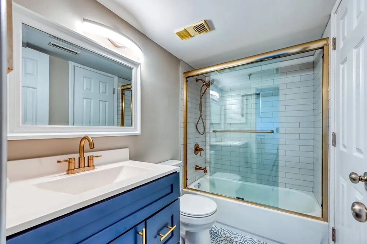 This modern bathroom features a vibrant blue vanity with gold fixtures, a glass-enclosed shower, and stylish patterned floor tiles. The overall look is fresh and contemporary, ideal for both comfort and function.