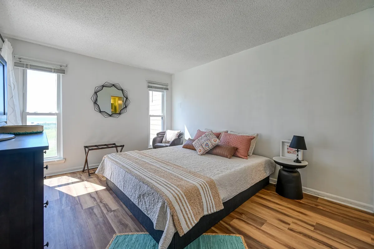 A serene primary bedroom with a peaceful mood, featuring a bed, decorative mirror, armchair, and small side table. Sunlight streams in, suggesting a calm retreat perfect for restful nights or quiet mornings, in keeping with the minimalist sanctuary theme.