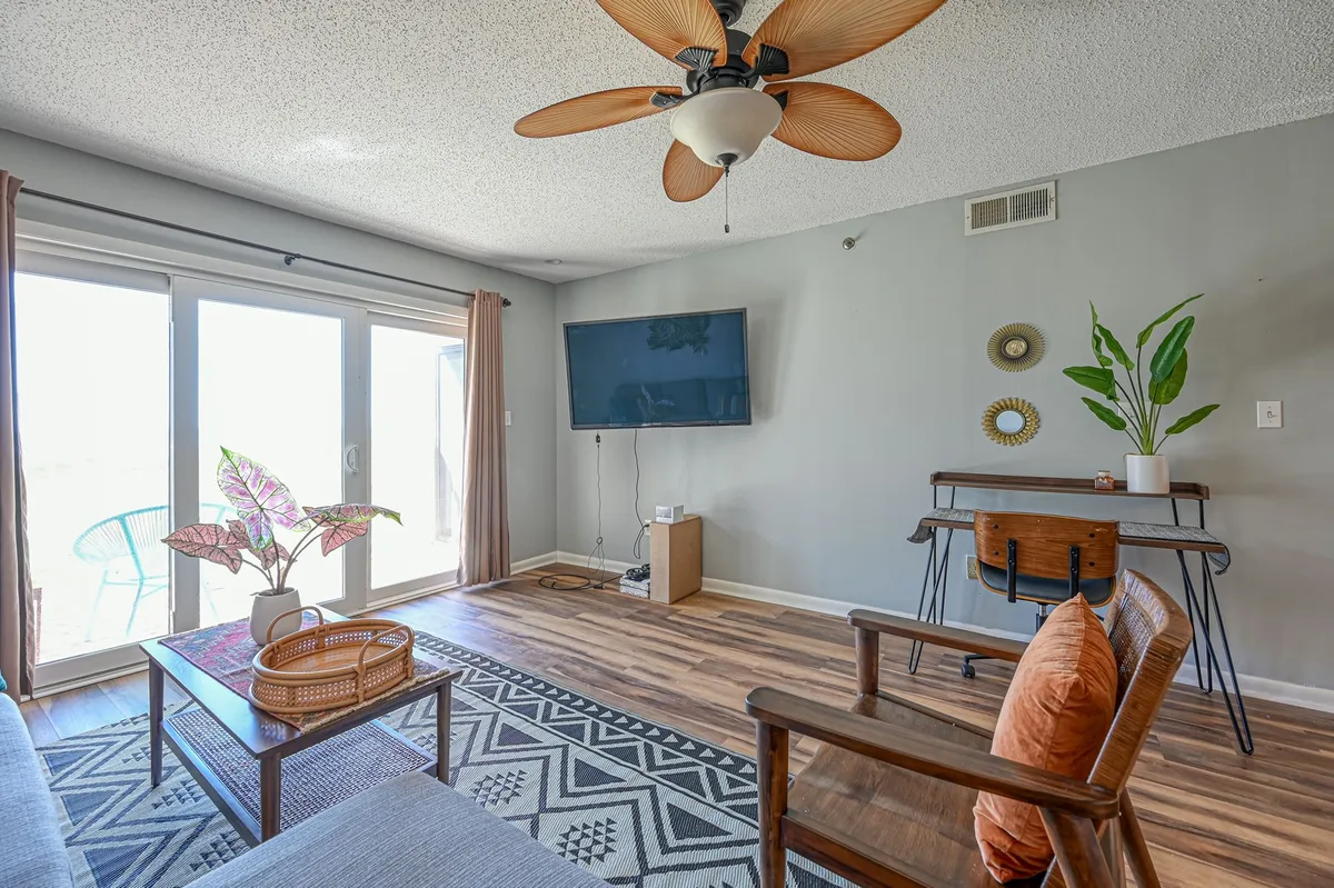 A bright, airy living area with large windows and sliding doors opening directly onto the balcony. The space features a stylish ceiling fan, colorful houseplant, modern TV, and decorative wall mirrors, creating a comfortable and inviting atmosphere ideal for relaxation while still being visually connected to the ocean.