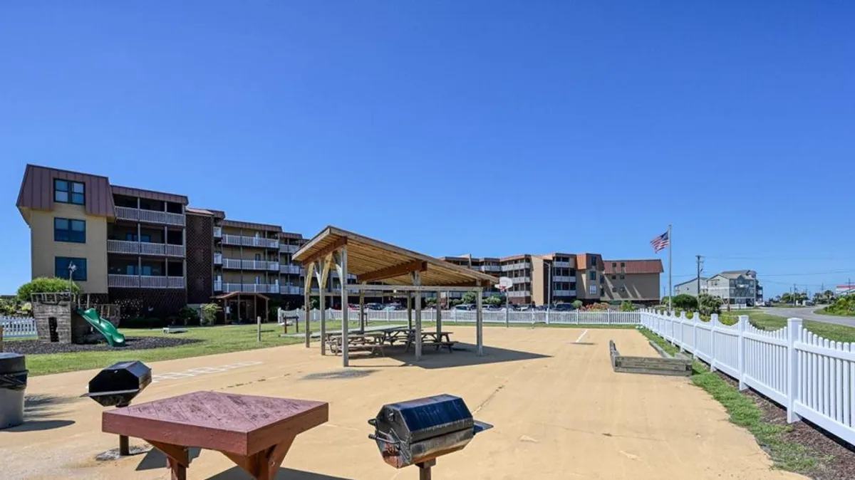 An inviting view of the resort's communal grilling area and picnic shelter, set against residential buildings. A playground slide and flagpole add to the welcoming, cheerful mood. The scene suggests a hub for sunset barbecues, reunions, and outdoor gatherings.