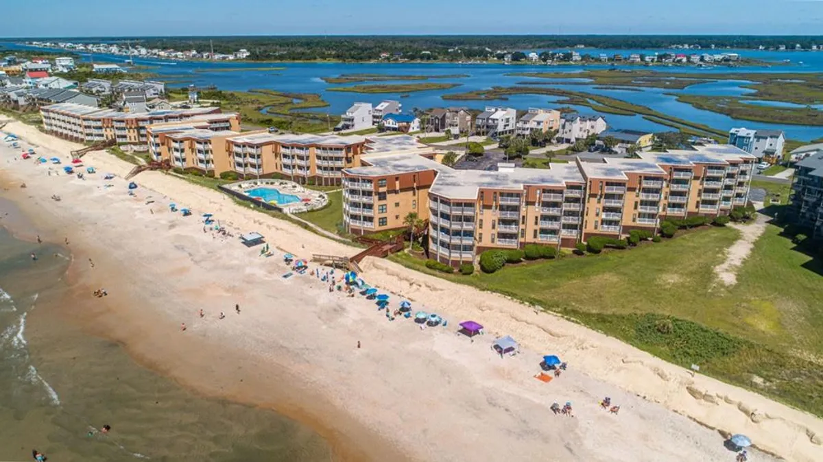 This vibrant exterior shot captures beachfront condos overlooking the swimming pool, with sunbathers, children playing beach games, families under umbrellas, and the ocean sparkling in the background. The mood is lively and carefree, with visible groups enjoying both the sand and the pool.