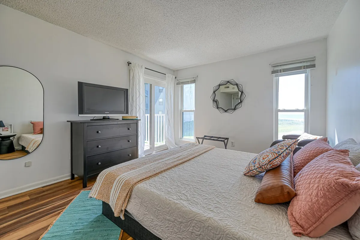 This image captures a serene bedroom featuring a neatly made bed with decorative pillows, a large mirror enhancing the sense of space, and a TV stand. Natural light filters gently into the room, creating a calm and inviting atmosphere ideal for restful nights.