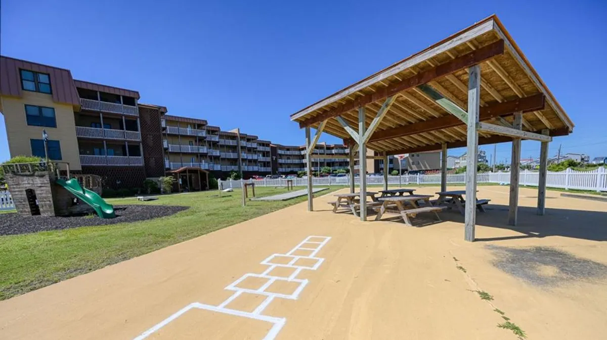A playful, sunlit exterior with a well-equipped playground, an inviting pavilion perfect for picnics, and a hopscotch court where children play. The image radiates a family-friendly, energetic mood, emphasizing spaces for games, gatherings, and childhood fun.