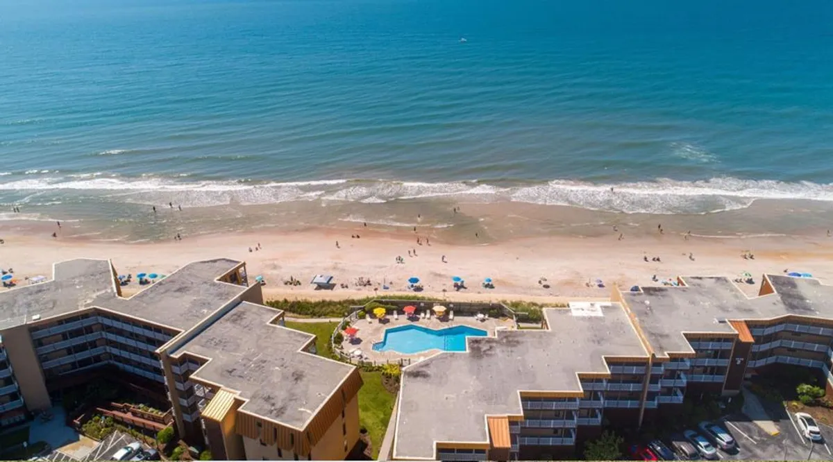 The hero image showcases an inviting oceanfront condo resort with direct beach access, a sparkling swimming pool surrounded by colorful umbrellas, and sweeping views of the Atlantic. The vibrant scene features guests enjoying poolside relaxation, beach volleyball, and sunbathing, all set against the backdrop of rolling ocean waves and soft sands.