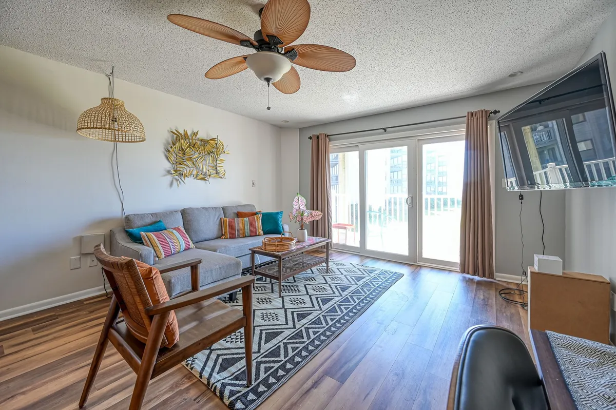 The living area features a comfortable sofa with colorful cushions, a coffee table, and large sliding glass doors that let in abundant natural light and reveal glimpses of the outdoors. The arrangement invites relaxation, conversation, or watching a movie with loved ones.