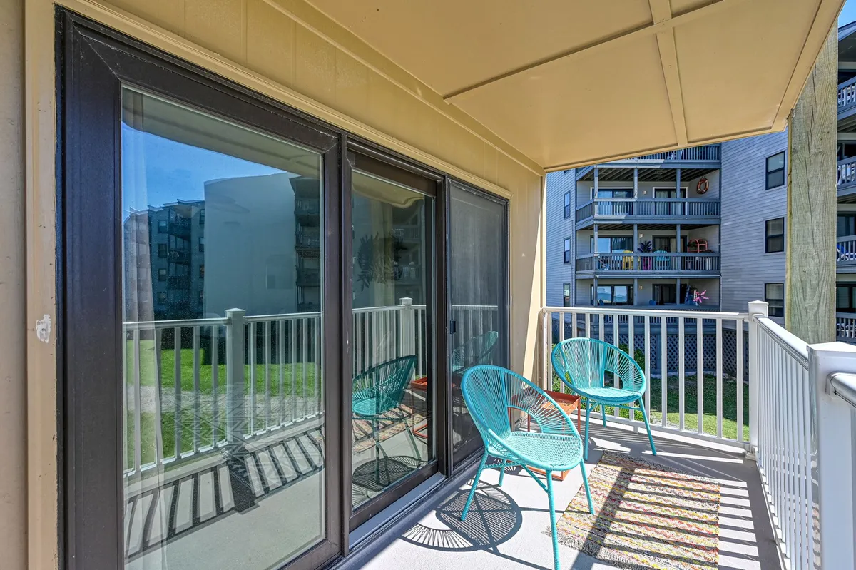 Two turquoise chairs set on a balcony or patio, positioned to face outward. Reflections in the glass doors hint at a cozy indoor space behind, while a lush green lawn is visible through the railing, blending indoor comfort with outdoor tranquility.