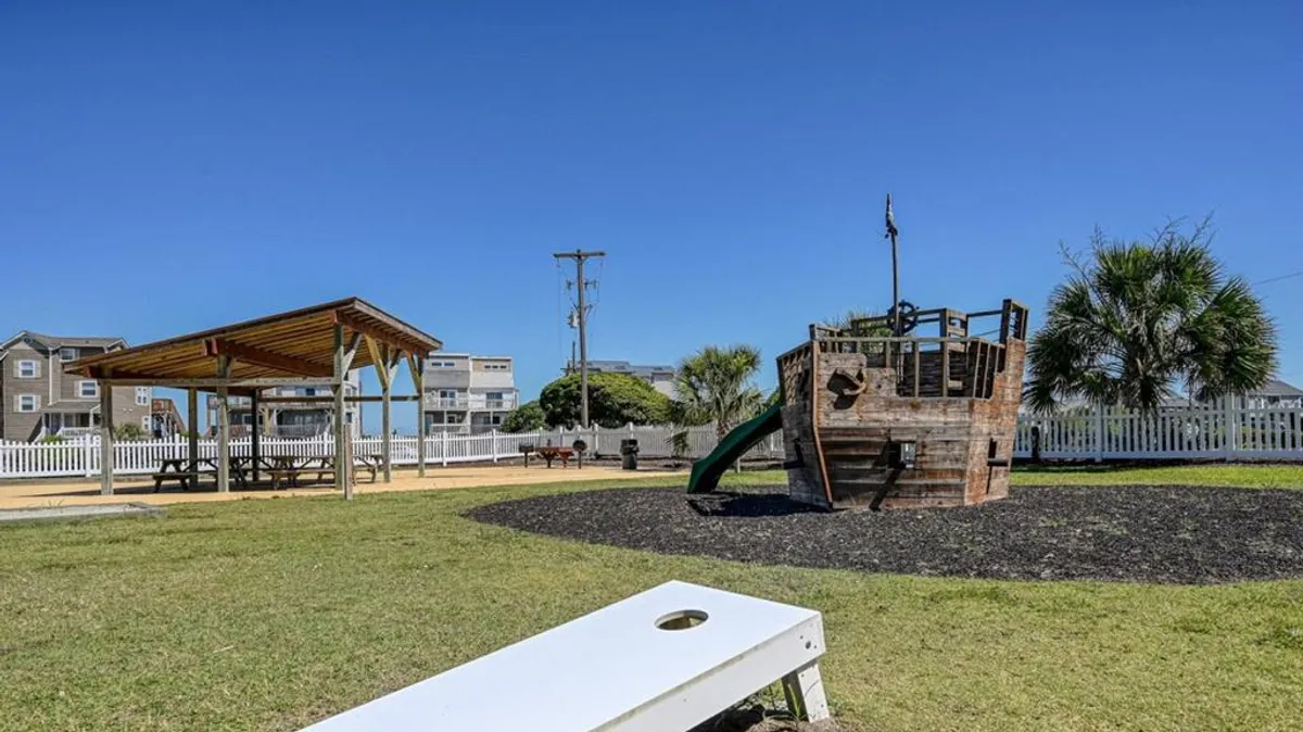 A playful exterior scene featuring a colorful playground designed as a pirate ship, with children climbing and playing. Lush green grass and a nearby picnic area invite families to gather and relax. The mood is energetic and joyful, underscoring community and family togetherness.